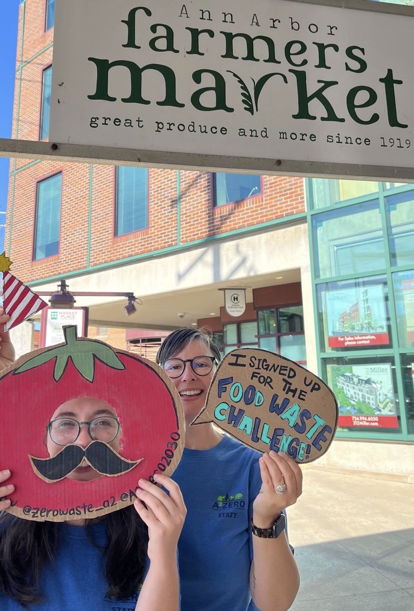 Two women in blue shirts hold cardboard cutouts of tomatoes with a speech bubble that says "I signed up for the Food Waste Challenge!"