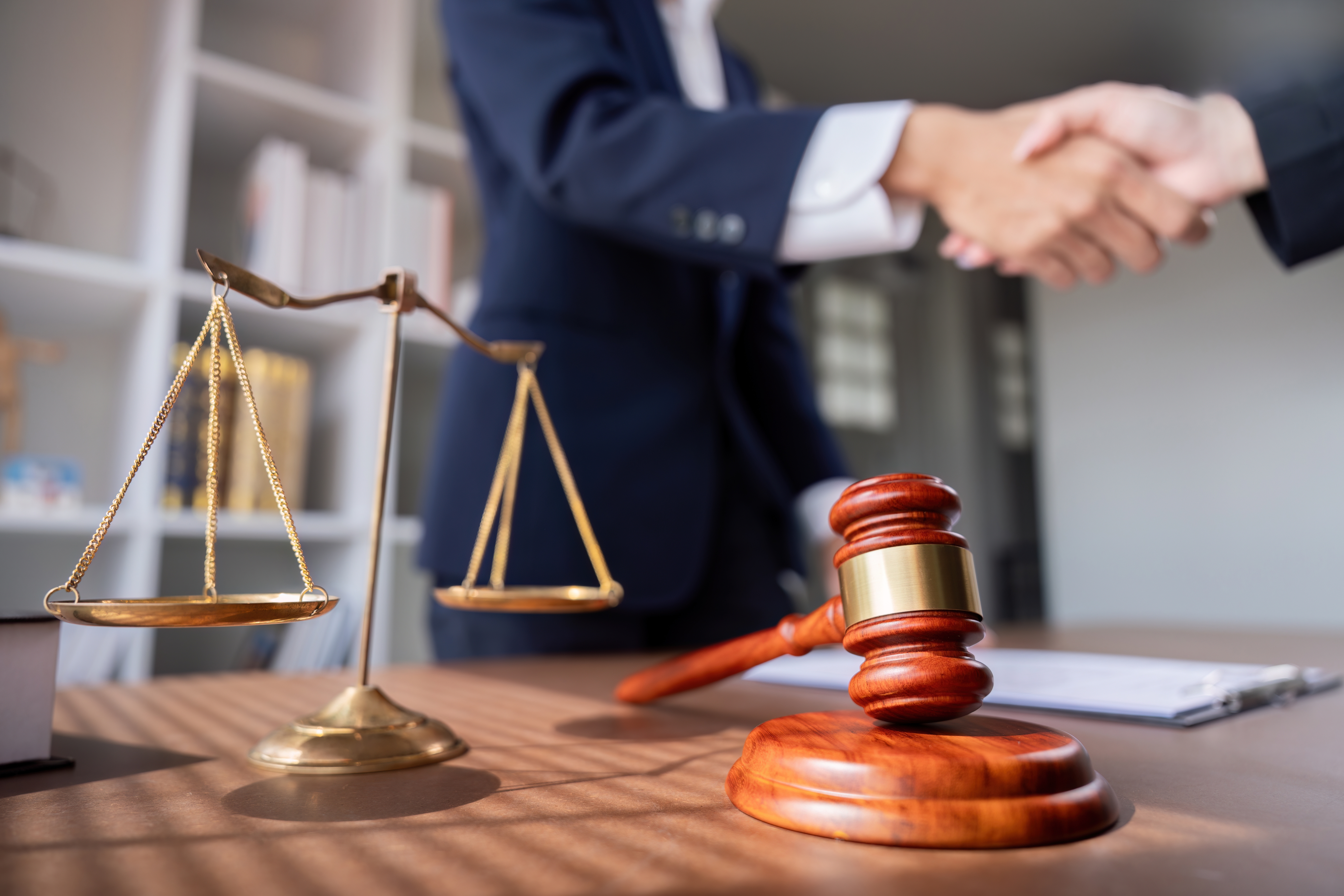 The scales of justice and a gavel sit on top of a desk. People in the background can be seen shaking hands. 