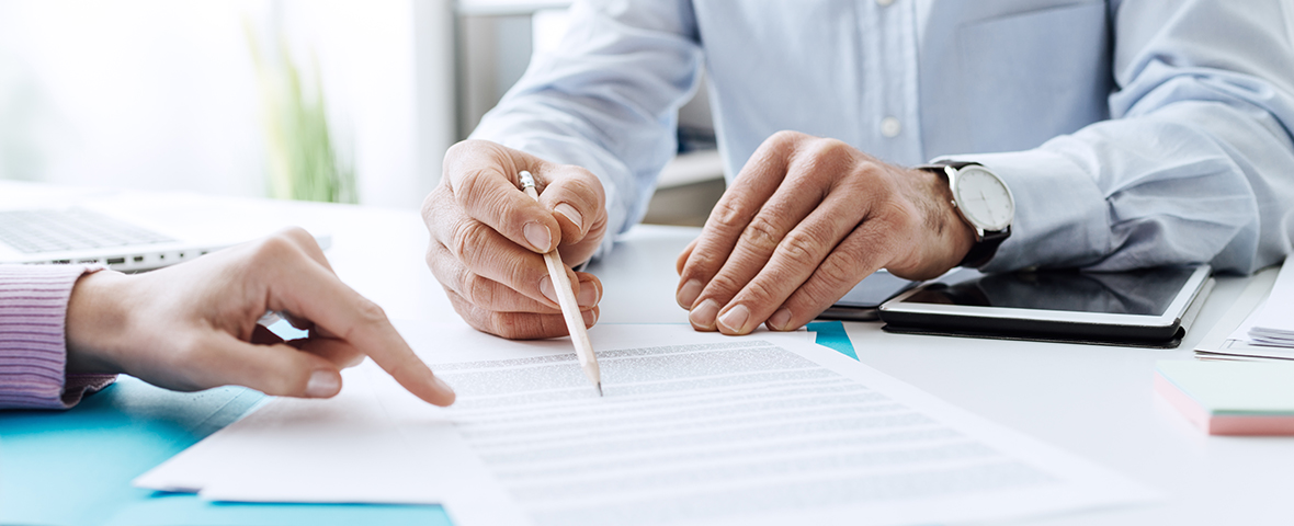 Two people sitting at a desk reviewing paperwork. The male on the right is holding a pencil to a piece of paper on the desk. The woman on the left is pointing to the same document on the table.